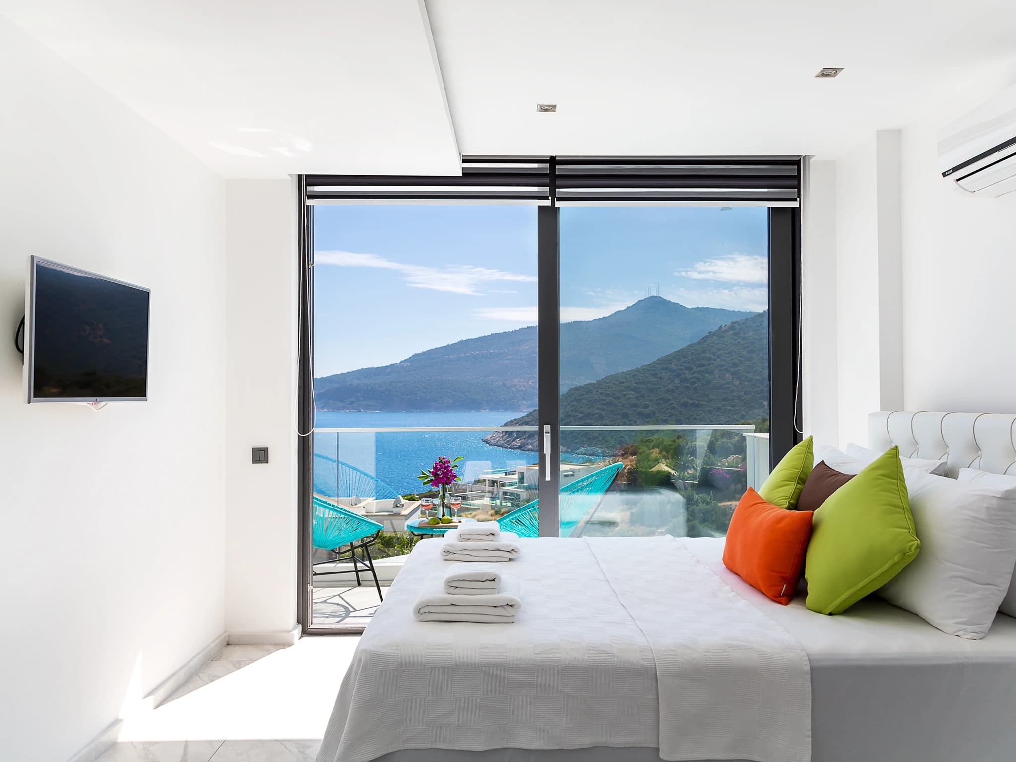 Bright modern bedroom with king bed, colorful accents, and expansive views to sea and mountains via glass doors.