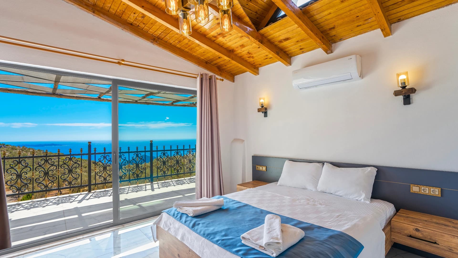 Bright bedroom with queen bed, wooden beams, and Mediterranean sea view from balcony.