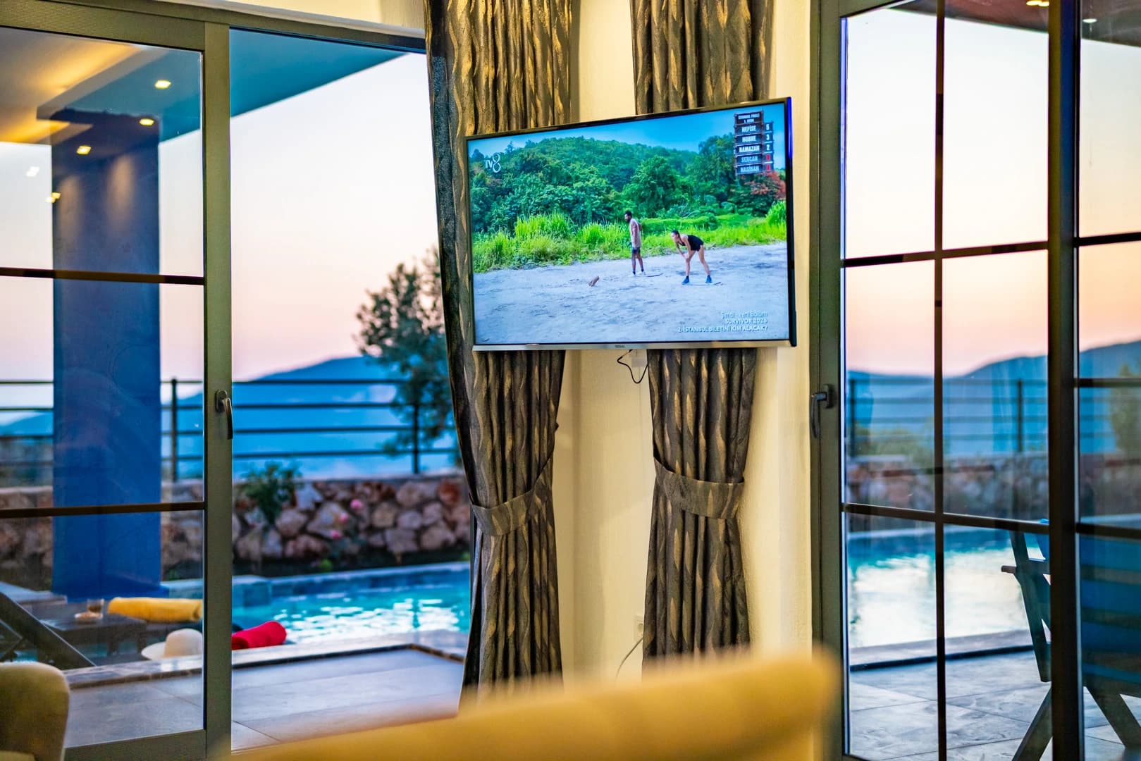 Living room with smart TV overlooking pool and scenic landscape through floor-to-ceiling windows.