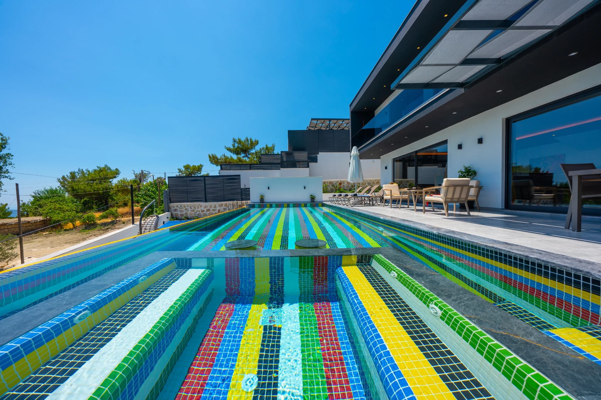 Vibrant lap pool with colorful mosaic tiles and lounging area beside the modern villa.