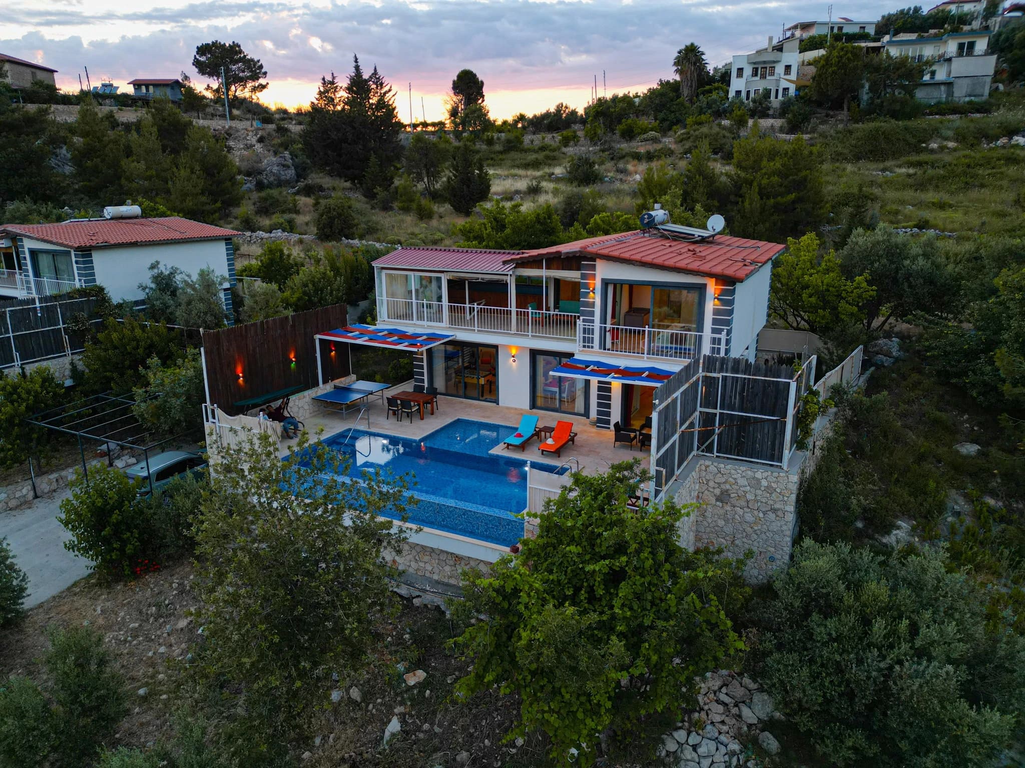 Abgeschiedene Villa mit Natur- und Bergblick, Innen- und Außenpool bei Kalkan – Aerial view of the villa at sunset with illuminated pool and colorful loungers.