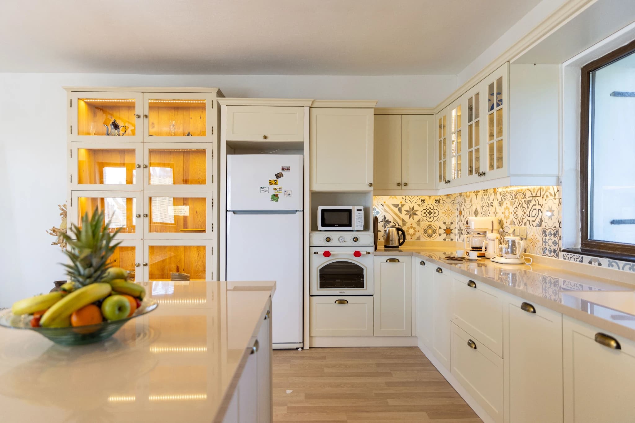 Modern cream-colored kitchen with integrated appliances, glass-door cabinets, and bright natural lighting.