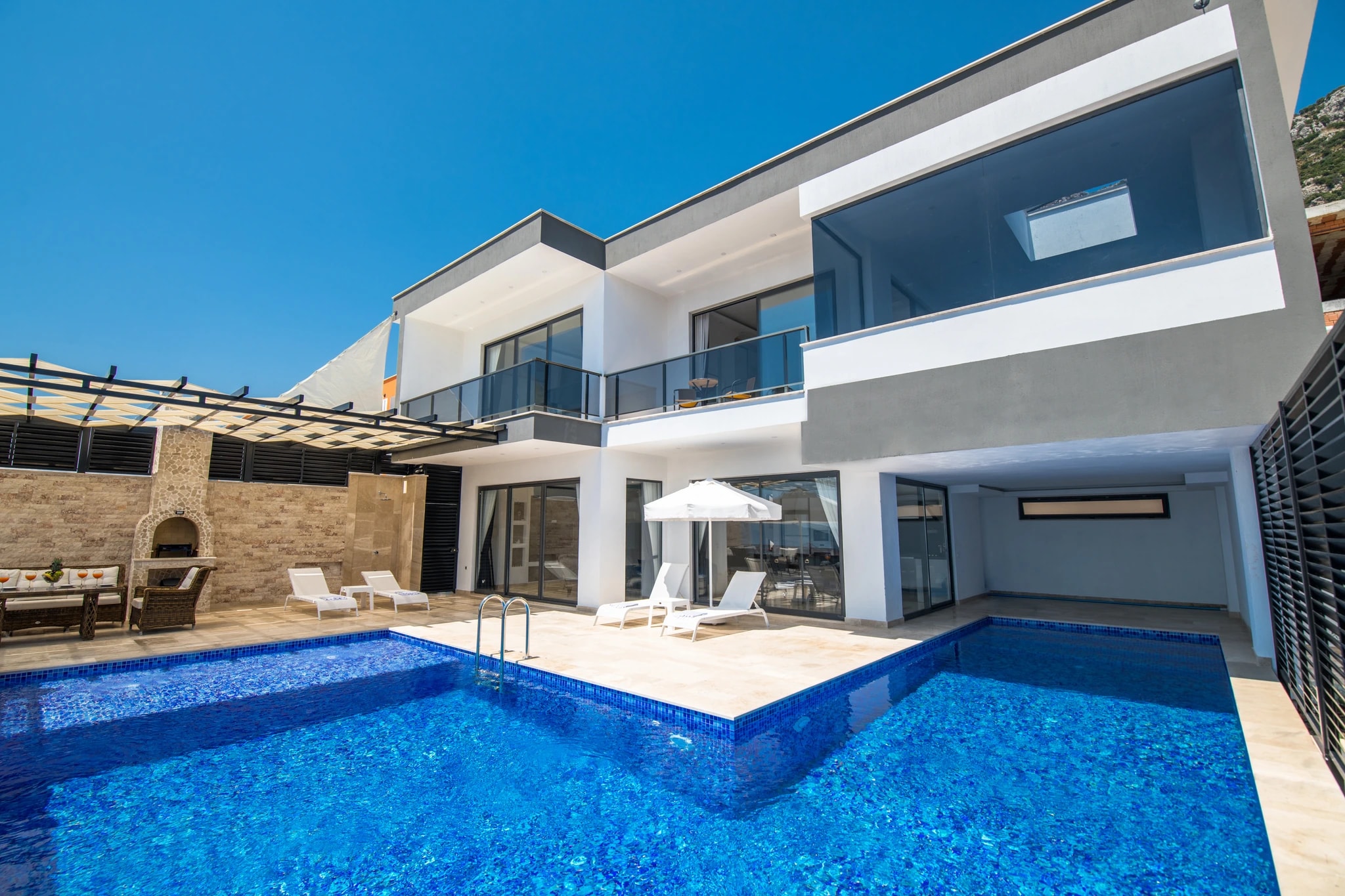 Secluded Honeymoon Villa with Private & Heated Indoor Pool in Kalkan – Modern villa exterior with large swimming pool, sun loungers, and outdoor seating area in bright daylight.