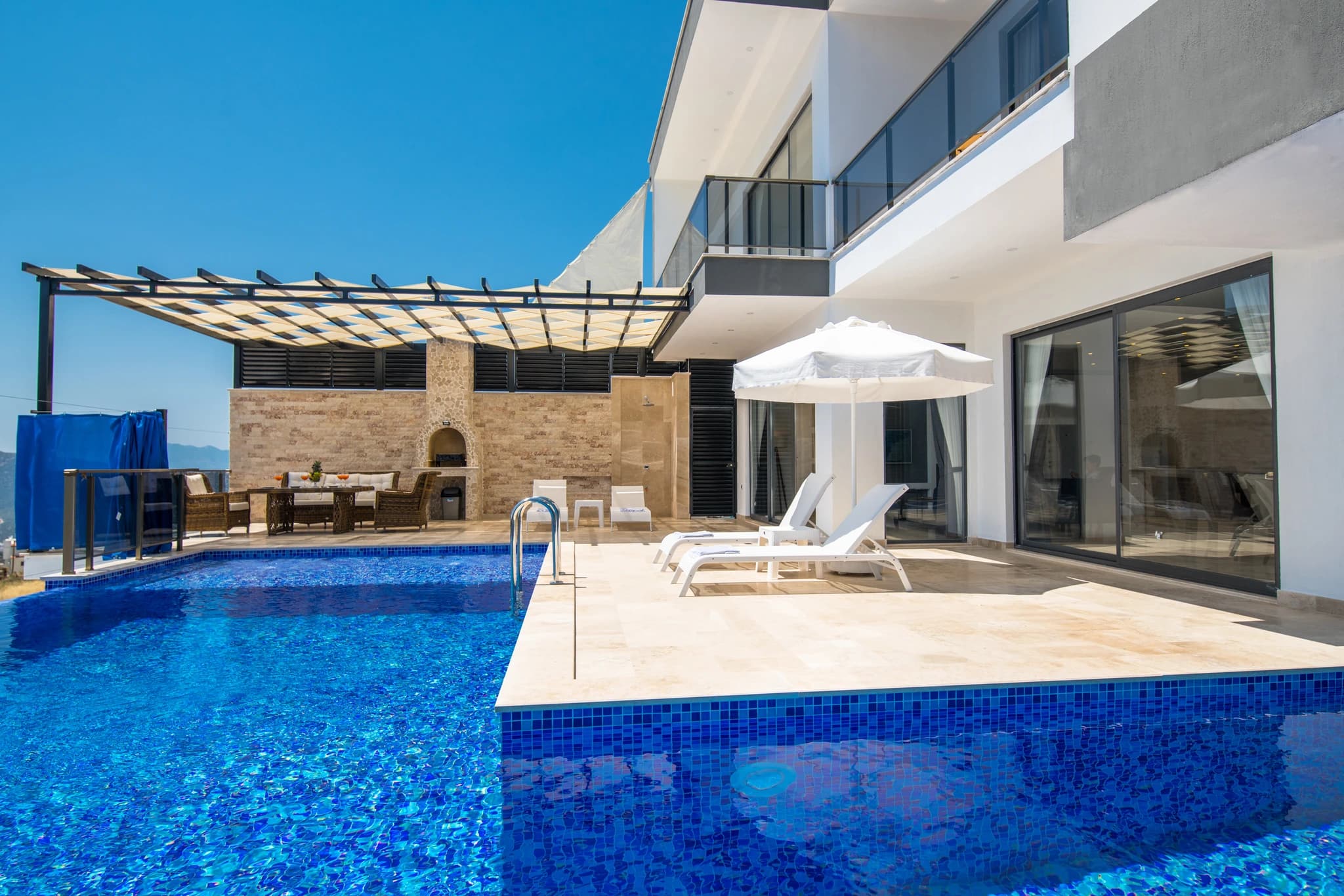 Secluded Honeymoon Villa with Private & Heated Indoor Pool in Kalkan – Resort-style pool area with dual pools, lounge chairs, pergola shade structure, and stone fireplace on expansive terrace.